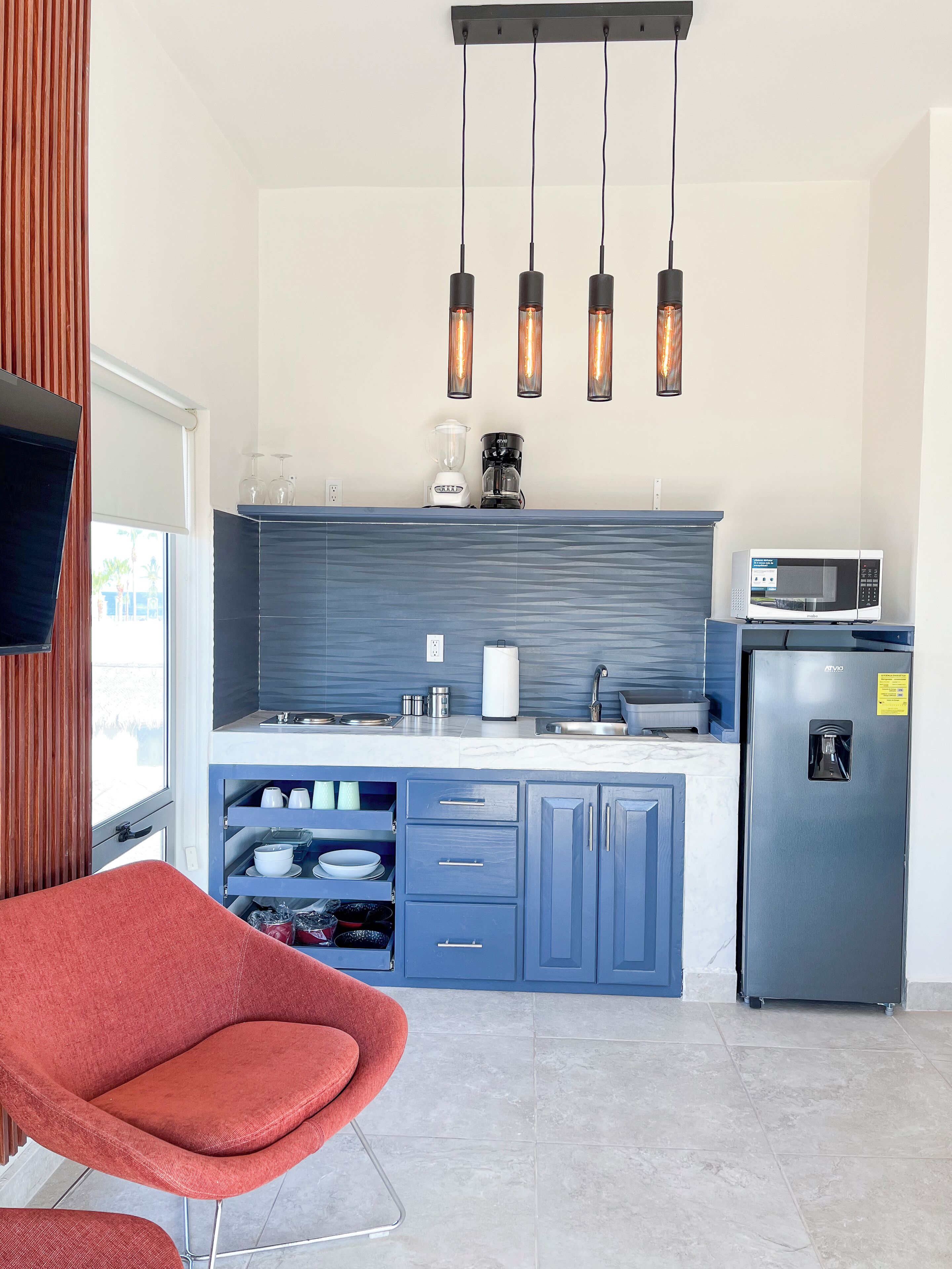A modern kitchen with blue cabinets, a grey refrigerator, a white microwave, and a red chair.
