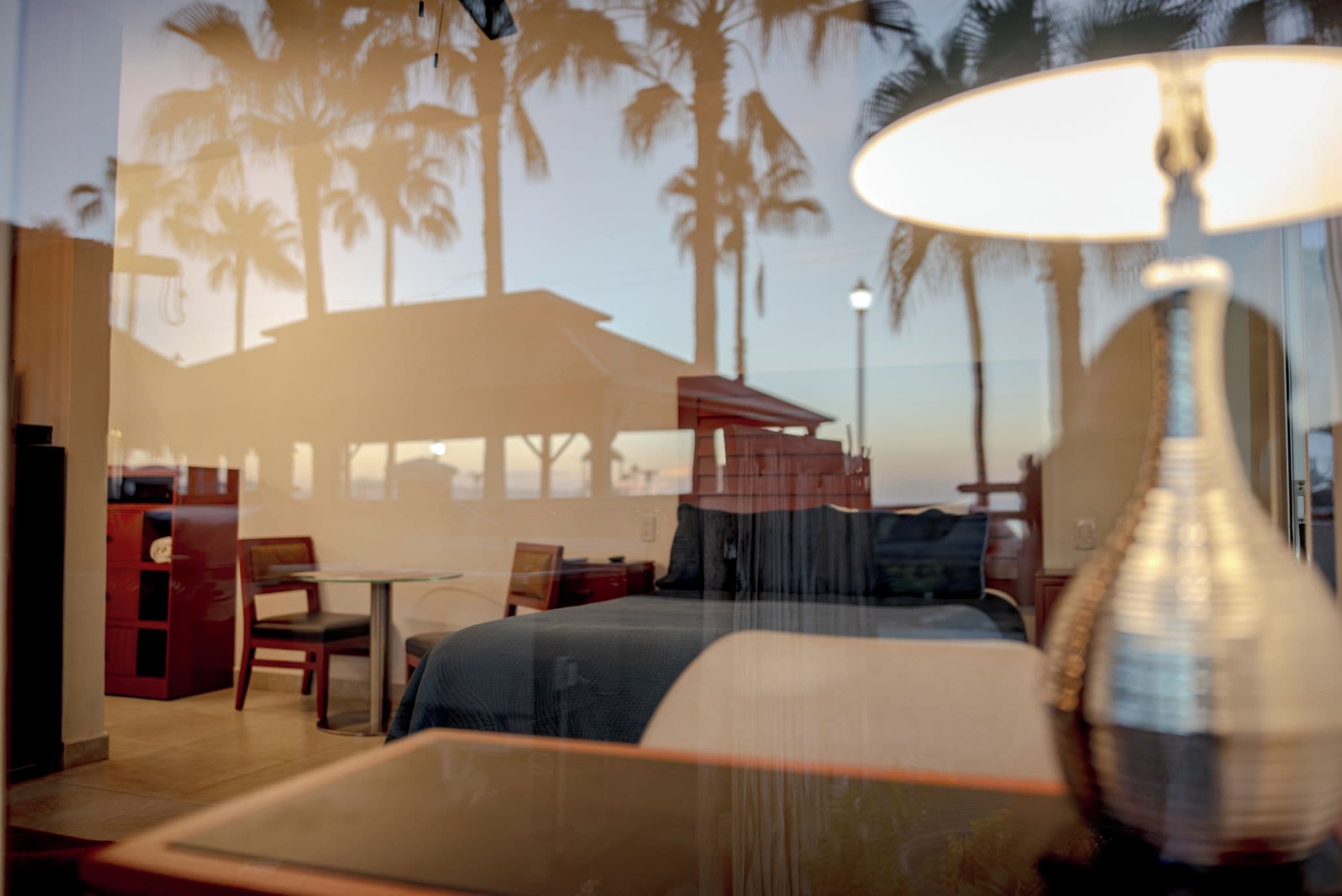 A room with a bed, a table, and chairs. There is a view of palm trees and a beach through the window.