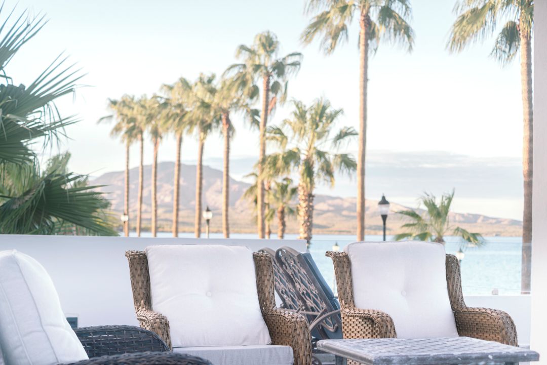 Wicker lounge chairs with white cushions, a small table, and a view of palm trees and a body of water.