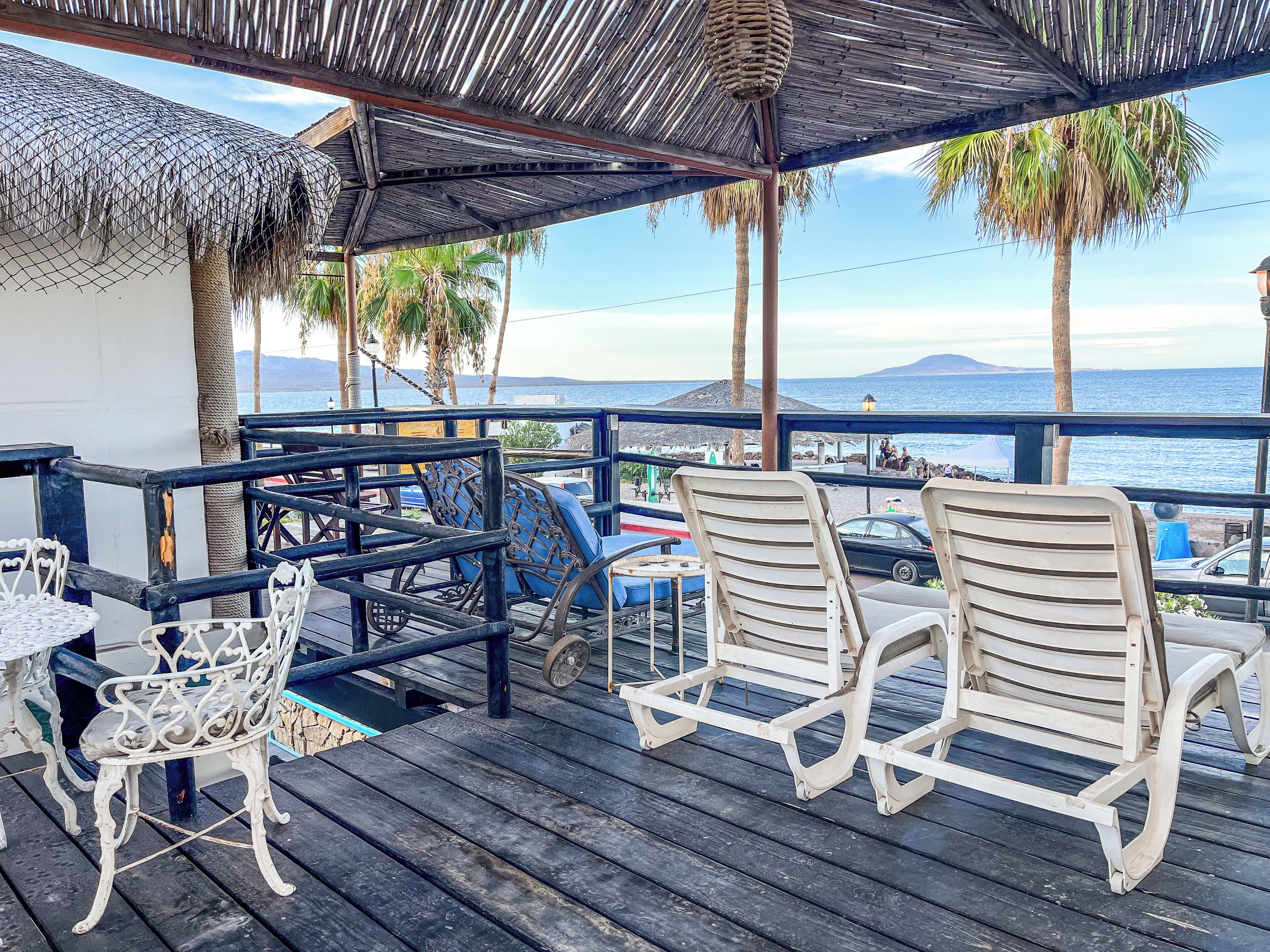 A wooden deck with chairs and tables, overlooking a beach with palm trees and the sea.