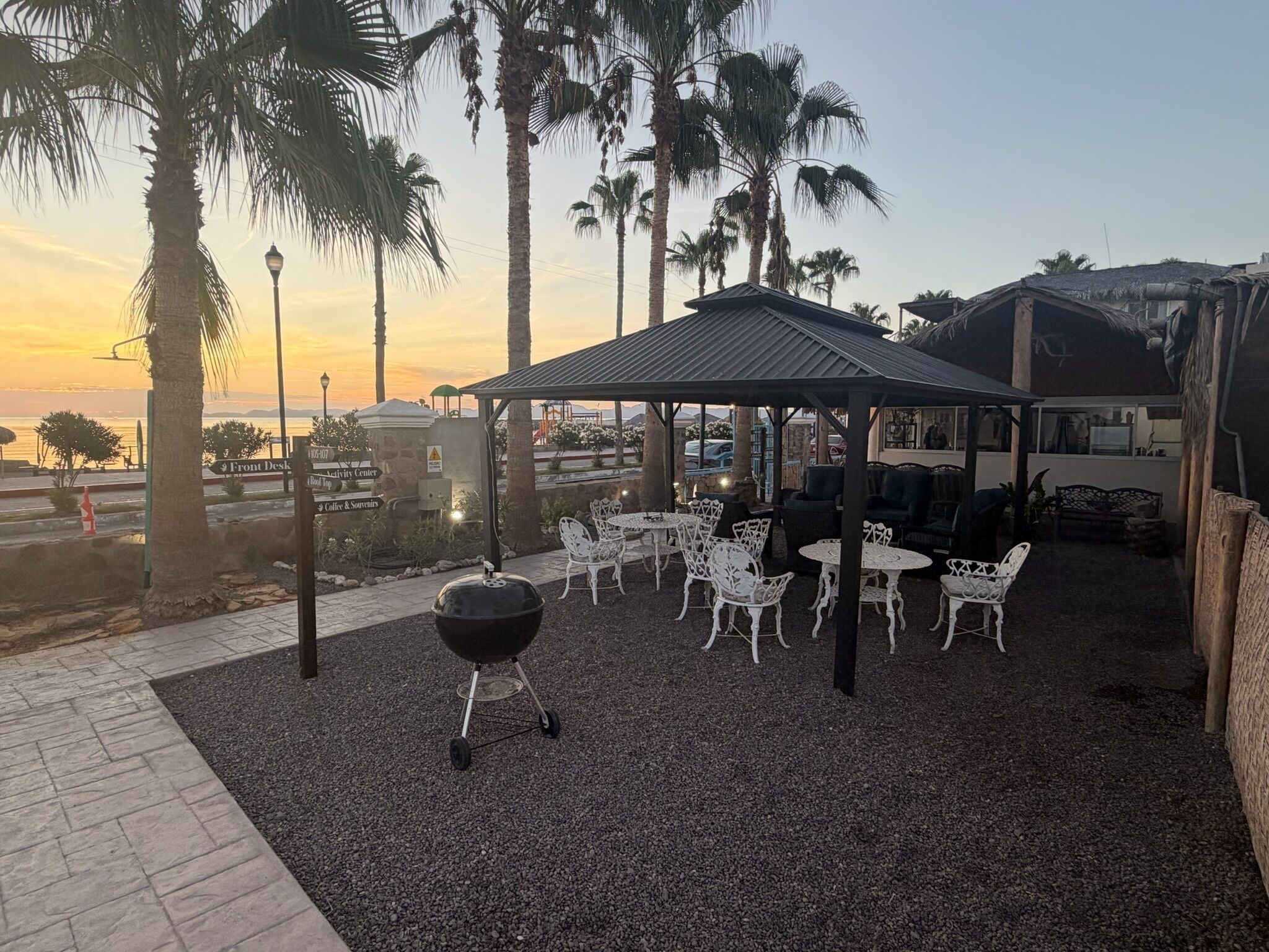 An outdoor patio area with a grill, white metal chairs, and tables, surrounded by palm trees and a view of the ocean at sunset.