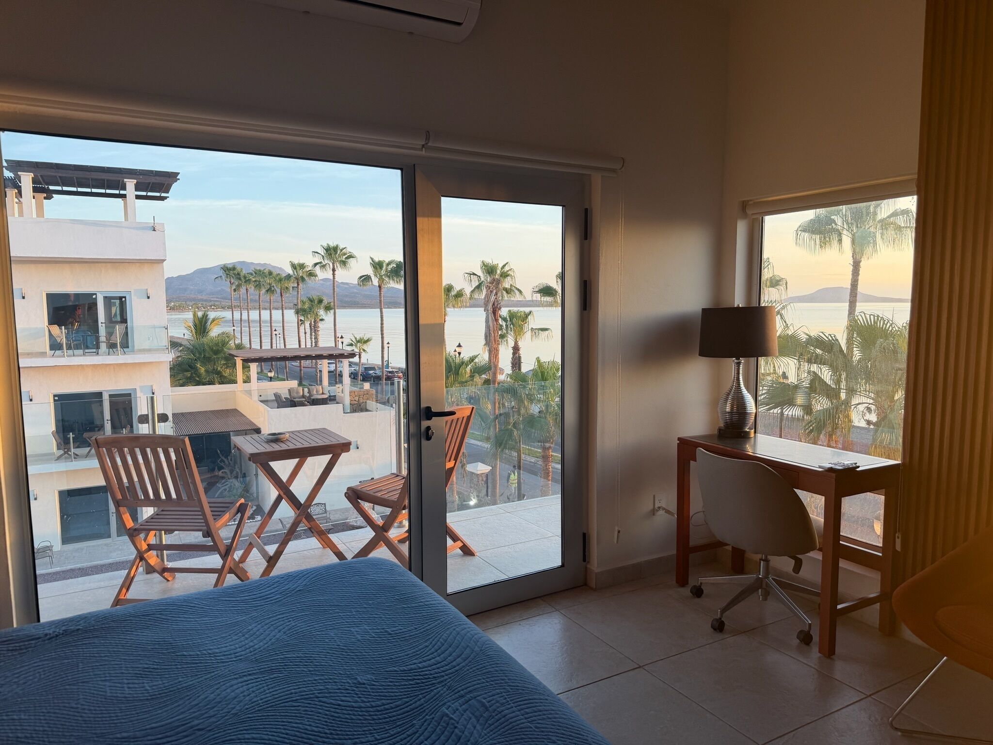 A hotel room with a bed, a desk, a chair, and a view of a pool and palm trees.