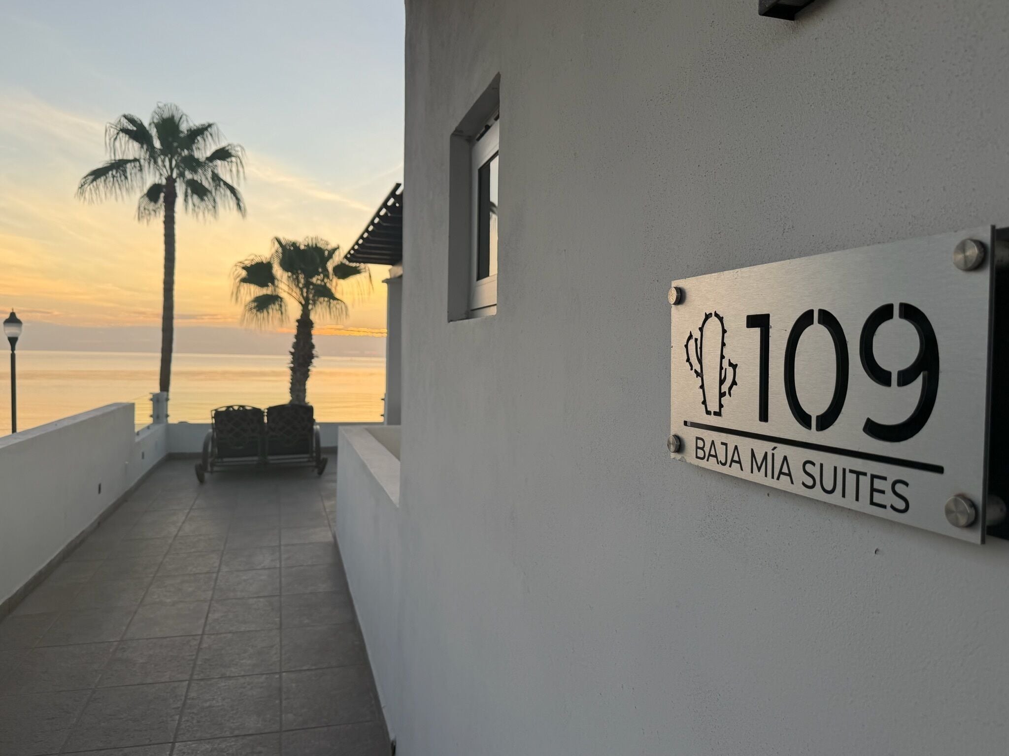 Ein Gebäude mit einem Schild, das '109 Baja Mía Suites' anzeigt, und Blick auf den Ozean mit Palmen.