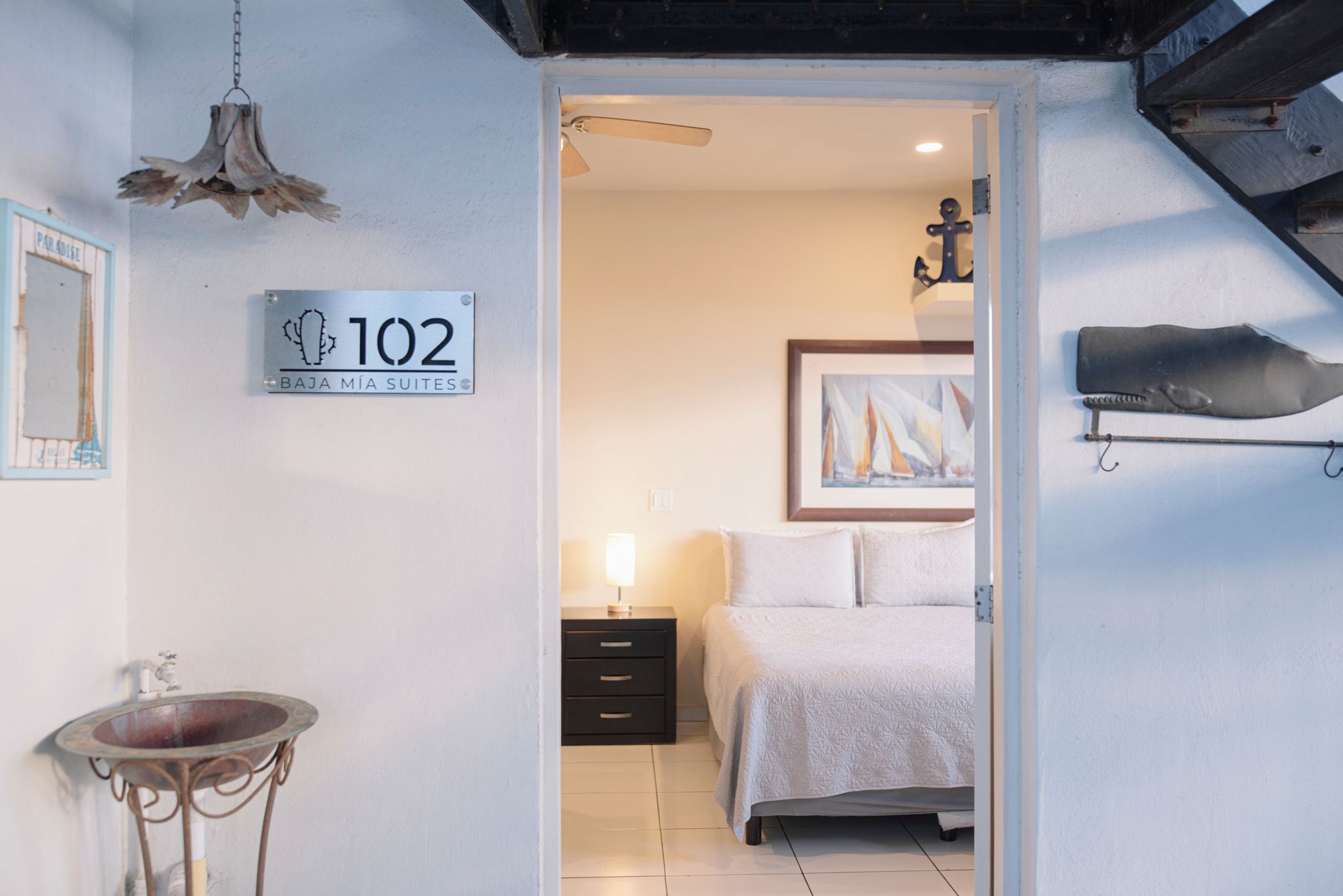 Quarto de hotel com cama, mesa de cabeceira, luminária, obra de arte na parede e placa indicando o número do quarto.