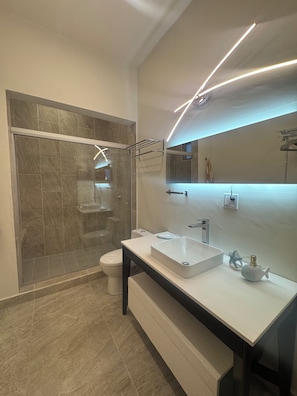 Un baño moderno con ducha de vidrio, lavamanos blanco y un espejo grande.