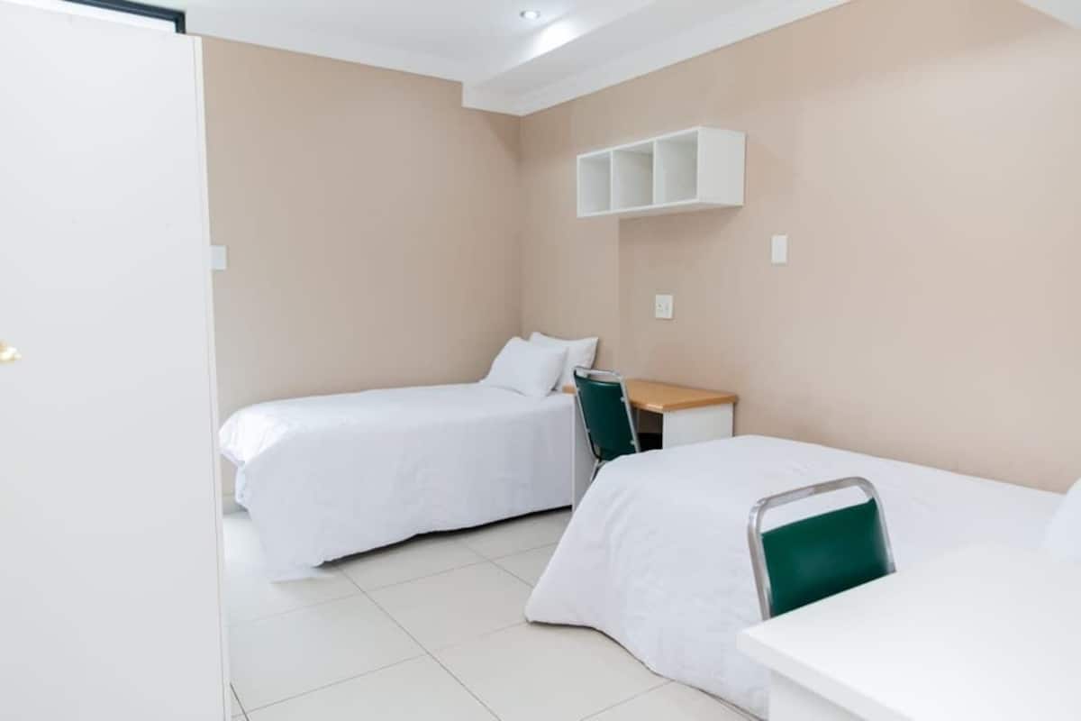 A small, clean room with two single beds, a desk, and a chair.