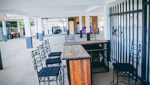 An indoor seating area with a long wooden table and metal chairs, a glass partition, and a view of the street outside. - Glenada Hotel (Johannesburg South)