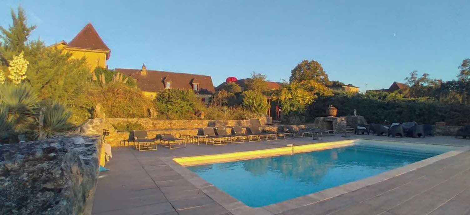 A swimming pool with lounge chairs, a stone wall, and a building with a red roof in the background.