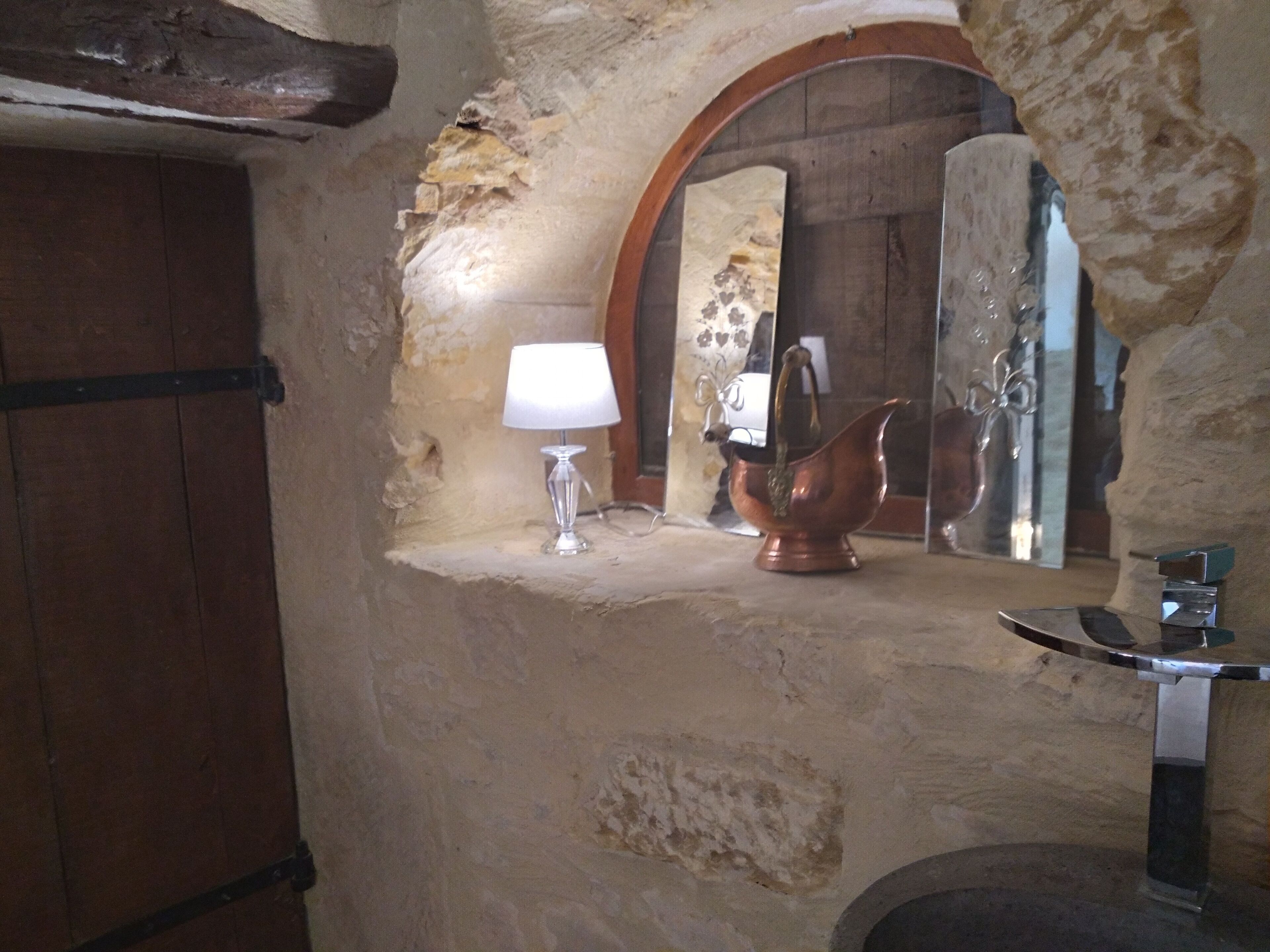 Une salle de bain rustique avec un mur en pierre, une porte en bois, une petite étagère sur laquelle reposent une lampe et un pichet en cuivre, et un miroir qui reflète une porte en bois et un motif floral.