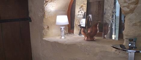 Une salle de bain rustique avec un mur en pierre, une porte en bois, une petite étagère sur laquelle reposent une lampe et un pichet en cuivre, et un miroir qui reflète une porte en bois et un motif floral.