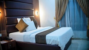 Une chambre d’hôtel avec un grand lit, une tête de lit en bois, une table de chevet avec un téléphone et une fenêtre avec des rideaux.