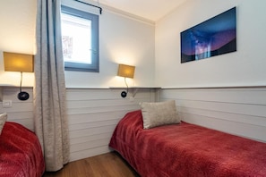 Une chambre avec deux lits, chacun recouvert d’une couverture rouge et agrémenté d’un oreiller. Deux lampes murales sont fixées de part et d’autre d’une fenêtre. Un tableau représentant une nuit étoilée est accroché au mur.