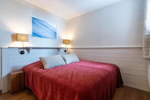 A neatly made bed with a red blanket, two bedside lamps, and a painting on the wall.