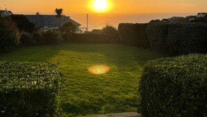 A grassy yard with a wooden deck, a hedge, and a view of the sunset over the ocean.