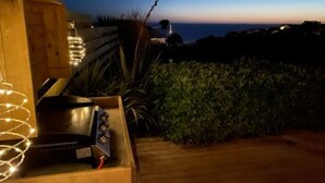 A wooden deck with a grill and string lights, overlooking a coastal landscape with a clear sky.