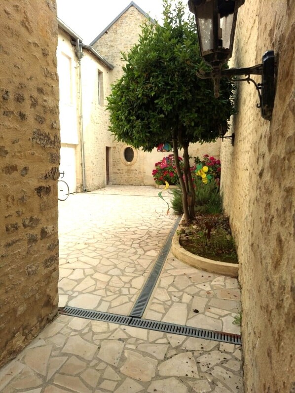 A narrow stone-paved courtyard with a tree and flower bed, surrounded by rustic stone walls.