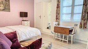 A bedroom with a bed, a desk, and a chair. There is a television on a stand, a door leading to another room, and a window with curtains. - Bayeux, Appartement Le Clem's'right in the heart of town and tourist sites. (Bayeux)