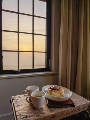 A table with a plate of food and two cups, positioned by a window with a view of a sunset.