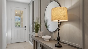 A hallway with a white door, a round mirror, a textured lamp, and two potted plants on a console table. - 6BR/5BA Luxurious with Private Pool (Kissimmee)