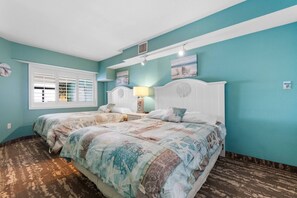 A bedroom with two beds, a window with shutters, a white headboard, and a wall-mounted lamp.