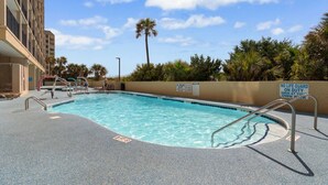 A swimming pool with a lifeguard sign, poolside chairs, and a palm tree in the background.
