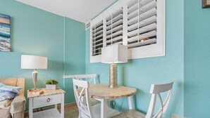 A room with teal walls, a round wooden table with chairs, a white lamp, and a window with shutters.
