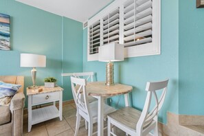 A room with teal walls, a round wooden table with chairs, a white lamp, and a window with shutters. - Cozy Oceanview at Beach Cove (North Myrtle Beach)