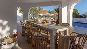 Un área de comedor al aire libre con mobiliario de mimbre, una mesa de madera y vistas a una piscina y estructuras con techo de paja.