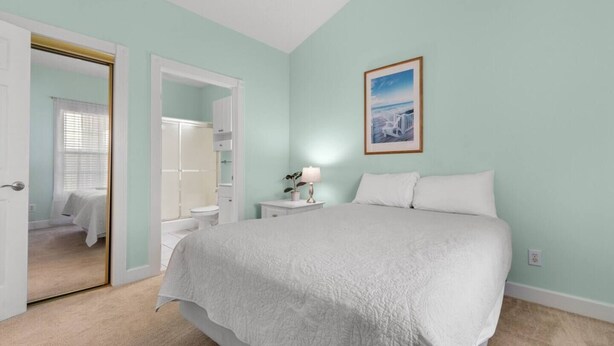 Quarto com cama, mesa de cabeceira com luminária, quadro pendurado na parede e porta que dá para outro cômodo.