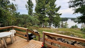 Una terraza de madera con una silla Adirondack roja y una mesa, con vista a un lago con árboles y un muelle al fondo.