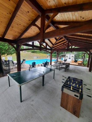 Un espacio recreativo al aire libre con piscina, mesa de fútbol y mesa de tenis bajo un pabellón de madera.