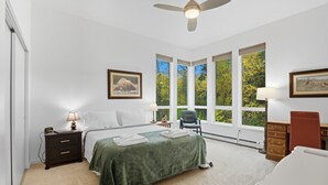 A bedroom with a bed, a desk, a chair, a nightstand, a lamp, a ceiling fan, and large windows with a view of trees. - Creekside 3BR Retreat Near Downtown, Airport & Fish Creek (Anchorage)