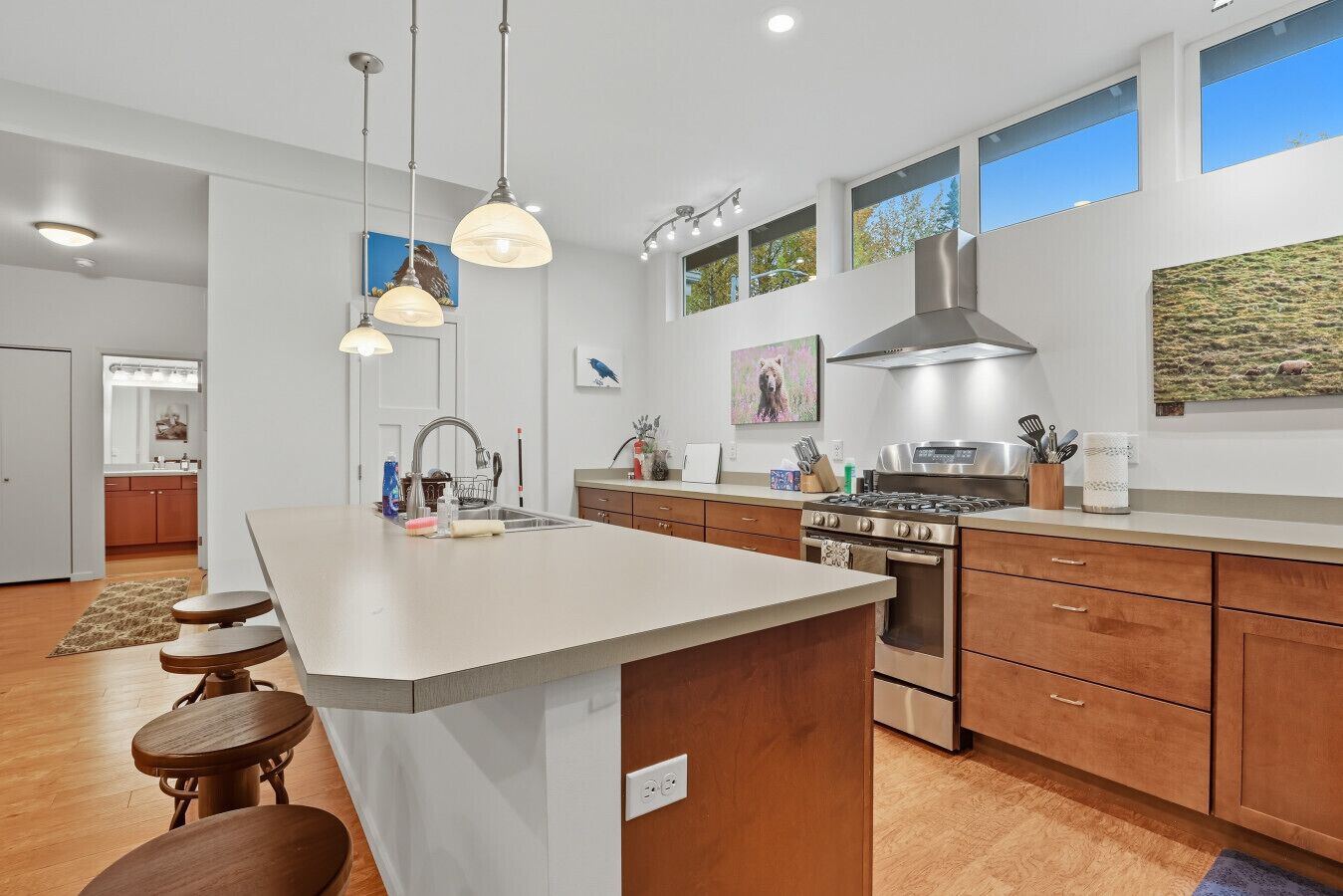 A modern kitchen with a central island, wooden cabinets, stainless steel appliances, pendant lighting, and artwork on the walls.