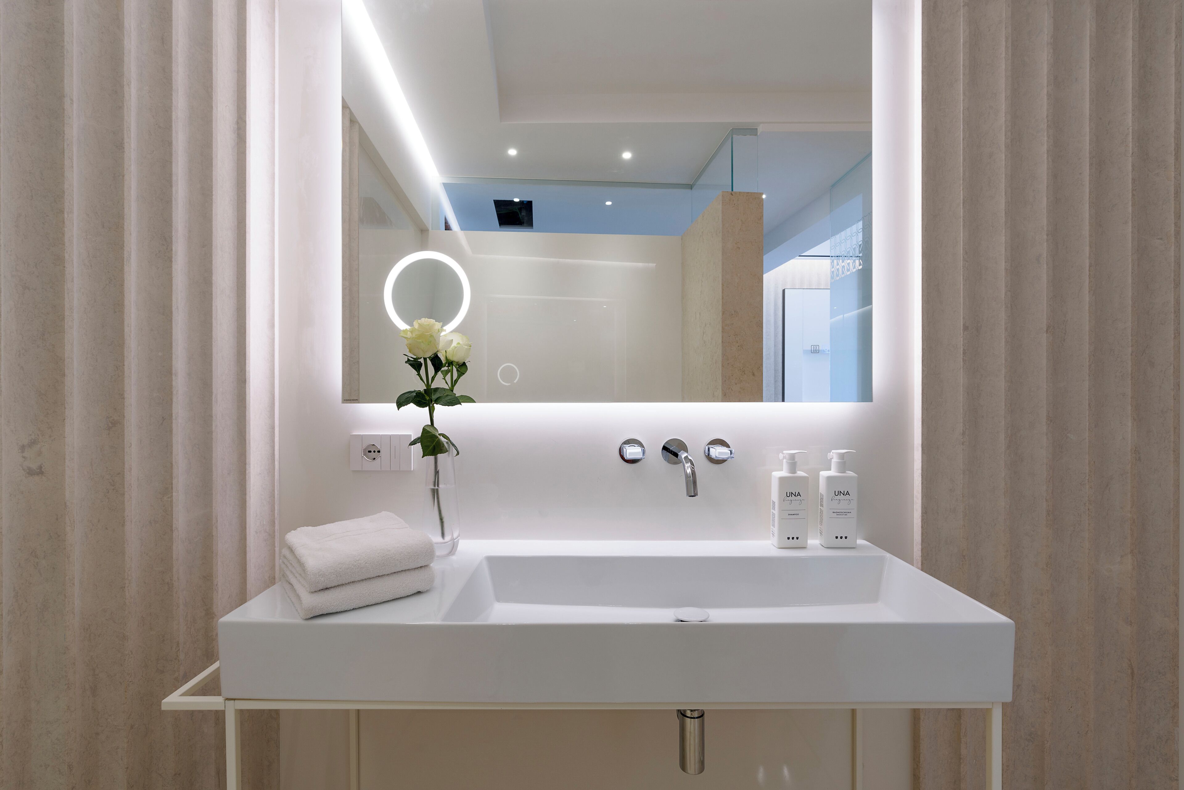 A modern bathroom with a white sink, a mirror with a circular light, and a vase with flowers.