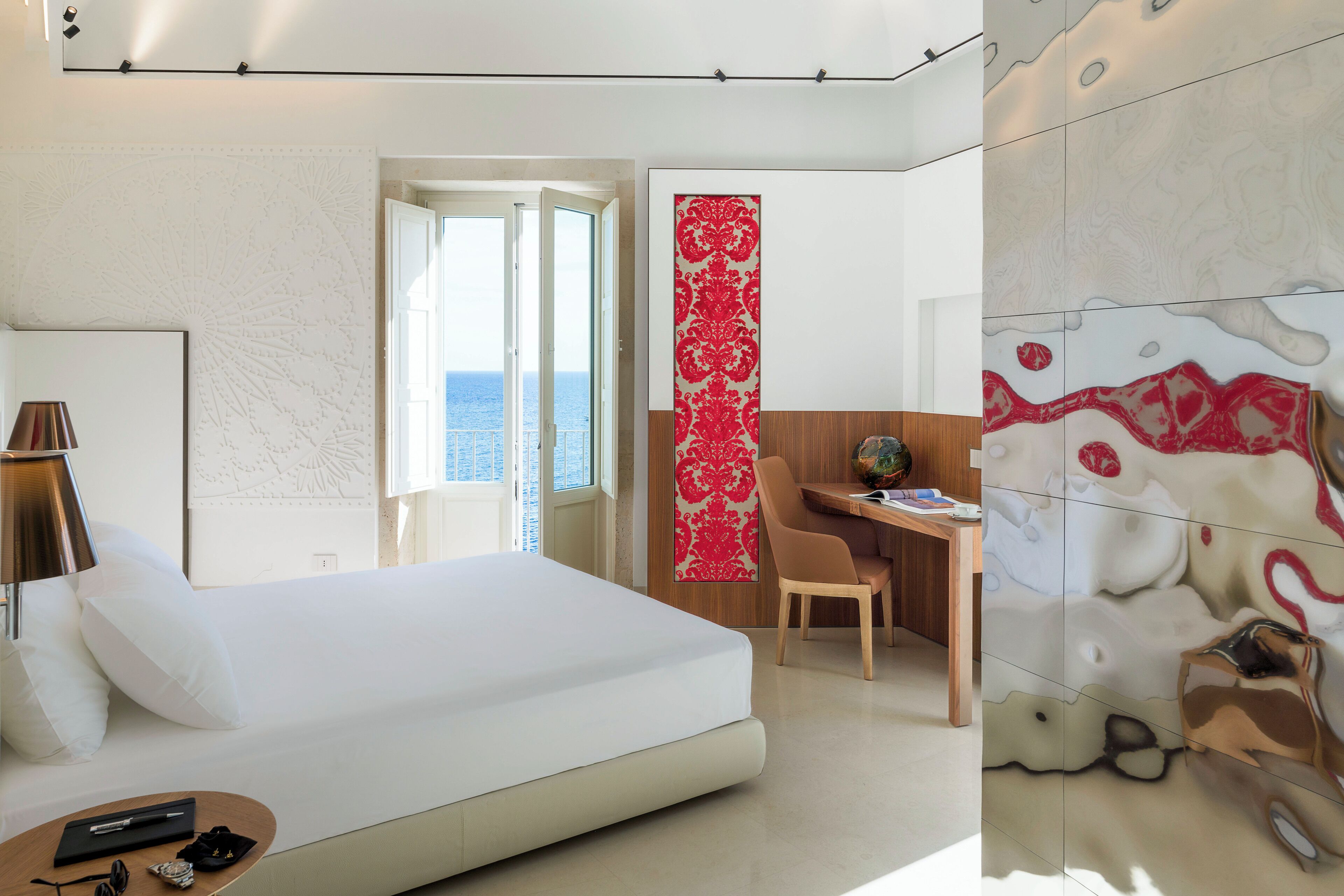 A modern hotel room with a large bed, a desk, and a view of the sea.