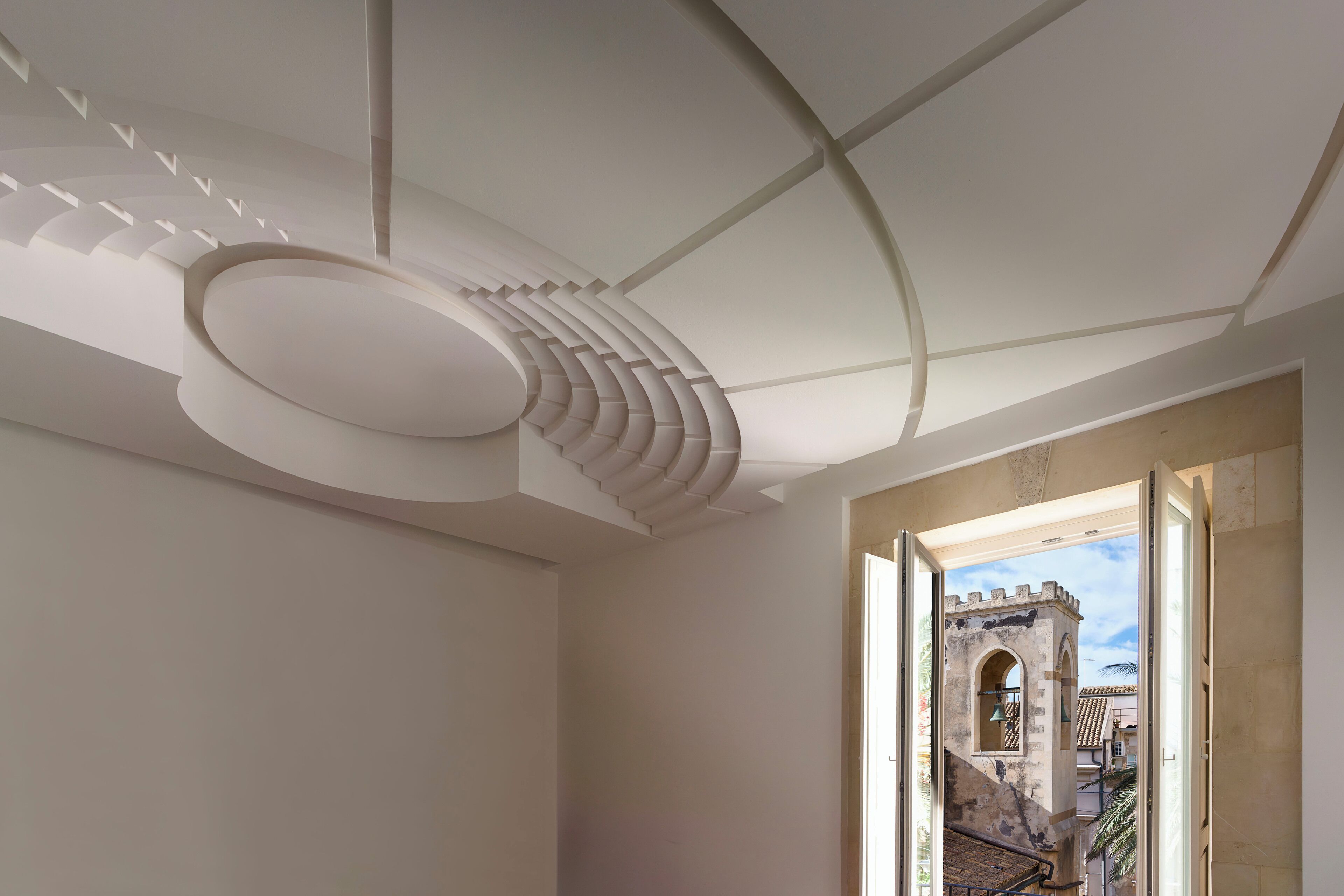 A room with a modern ceiling design, a view of a historic building through an open door, and a staircase leading outside.