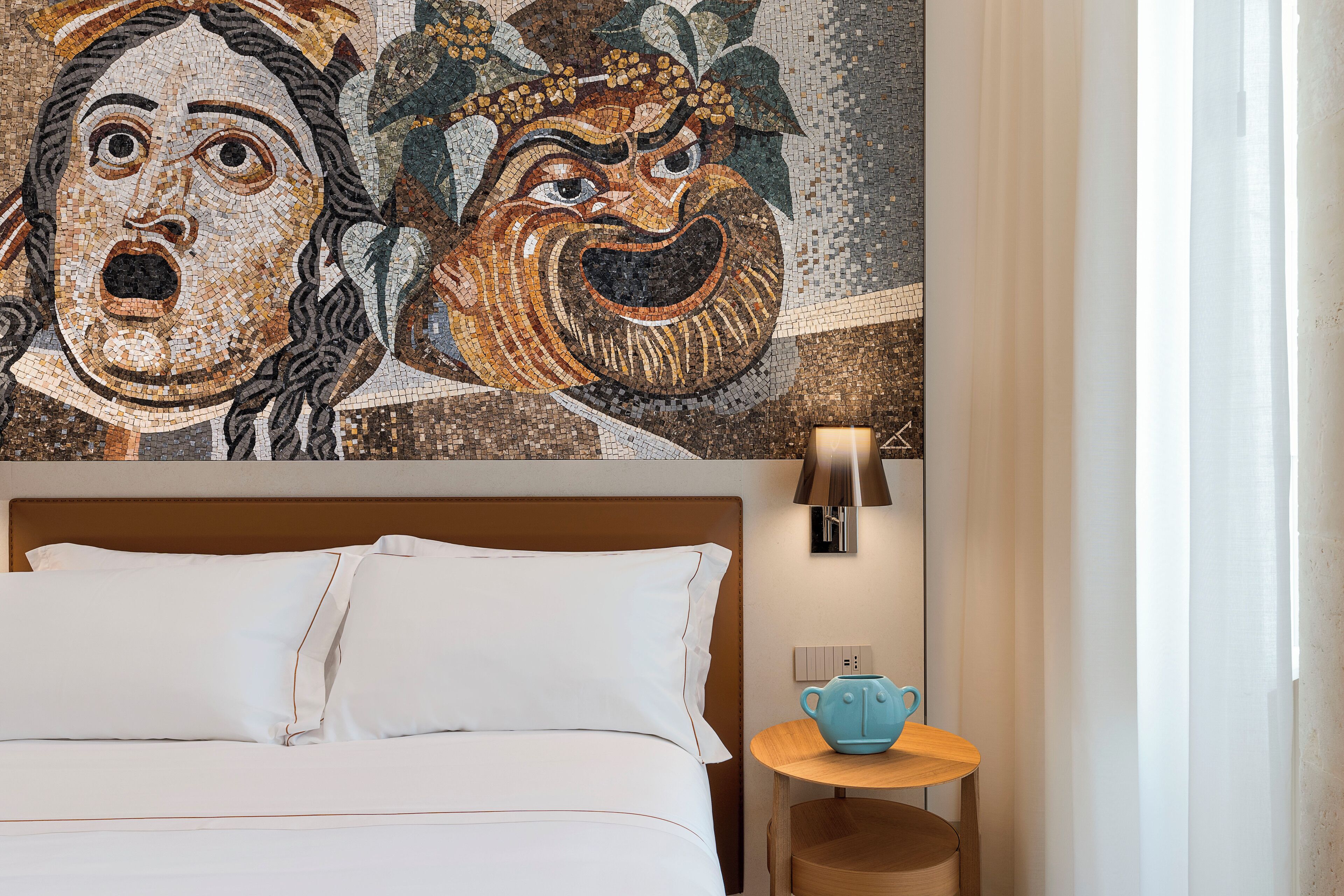 A hotel room with a bed, a nightstand with a lamp, and a wall mural.