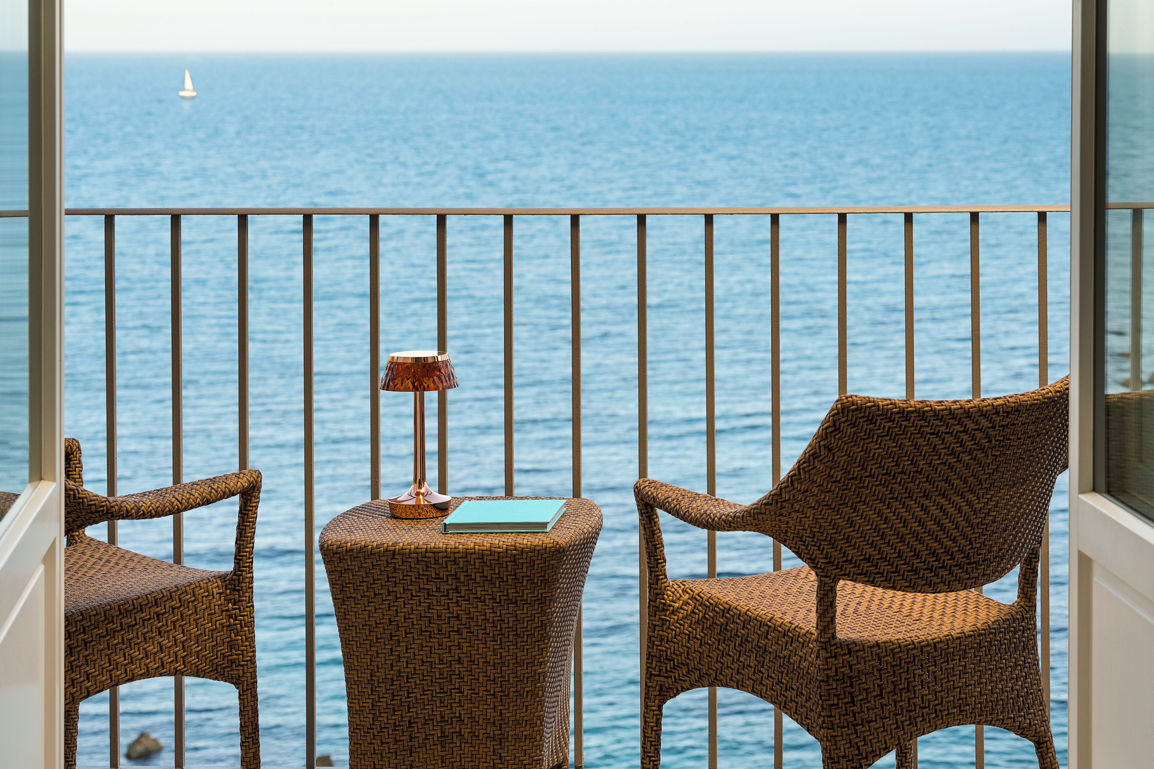 Un balcone con sedie in vimini e un tavolino, affacciato su uno specchio d'acqua con una barca a vela in lontananza.