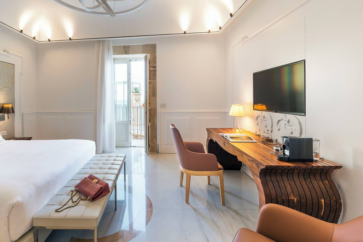 A hotel room with a bed, a bench, a desk with a chair, a TV, and a coffee machine.