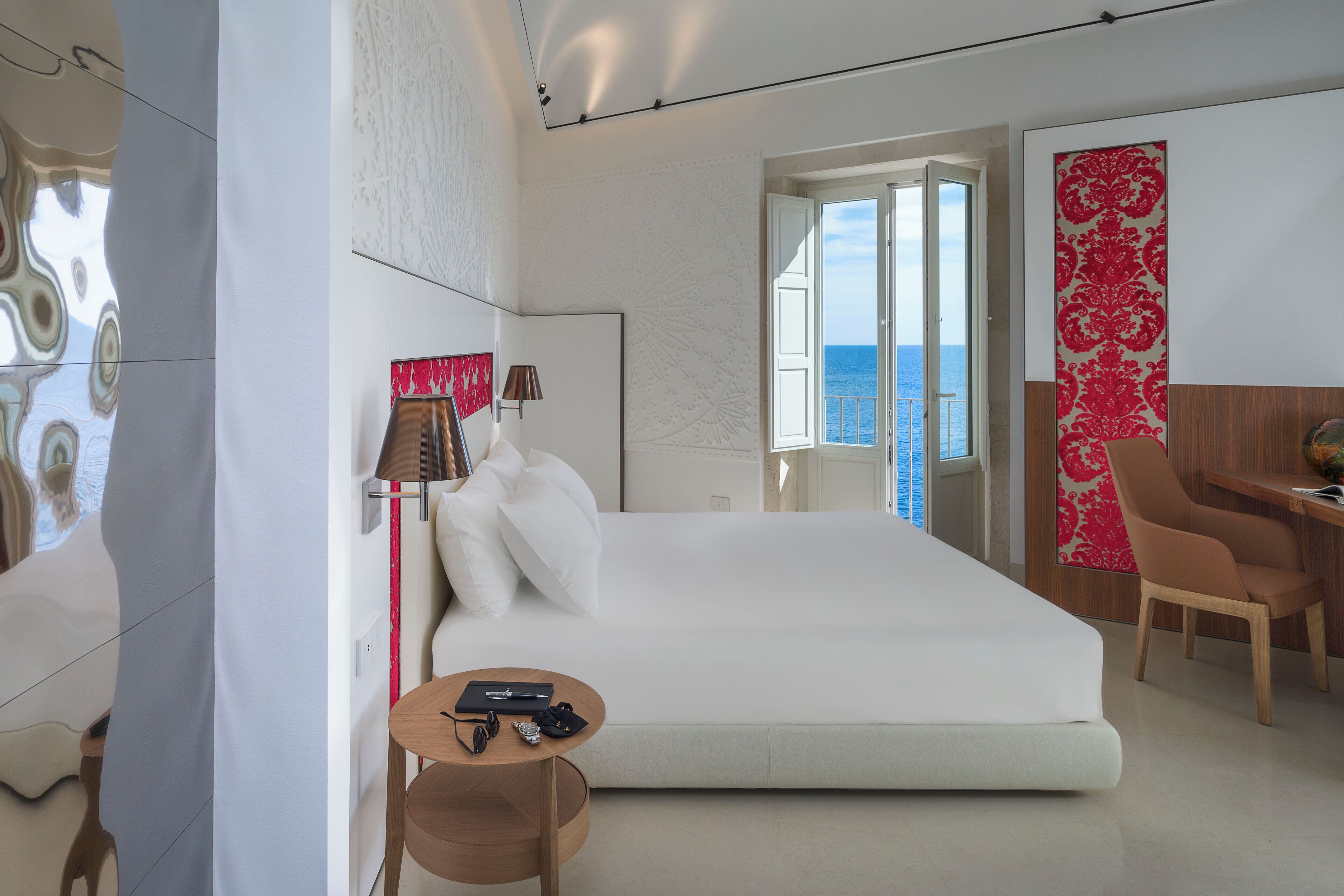 A modern hotel room with a large bed, a desk, a chair, and a view of the sea.