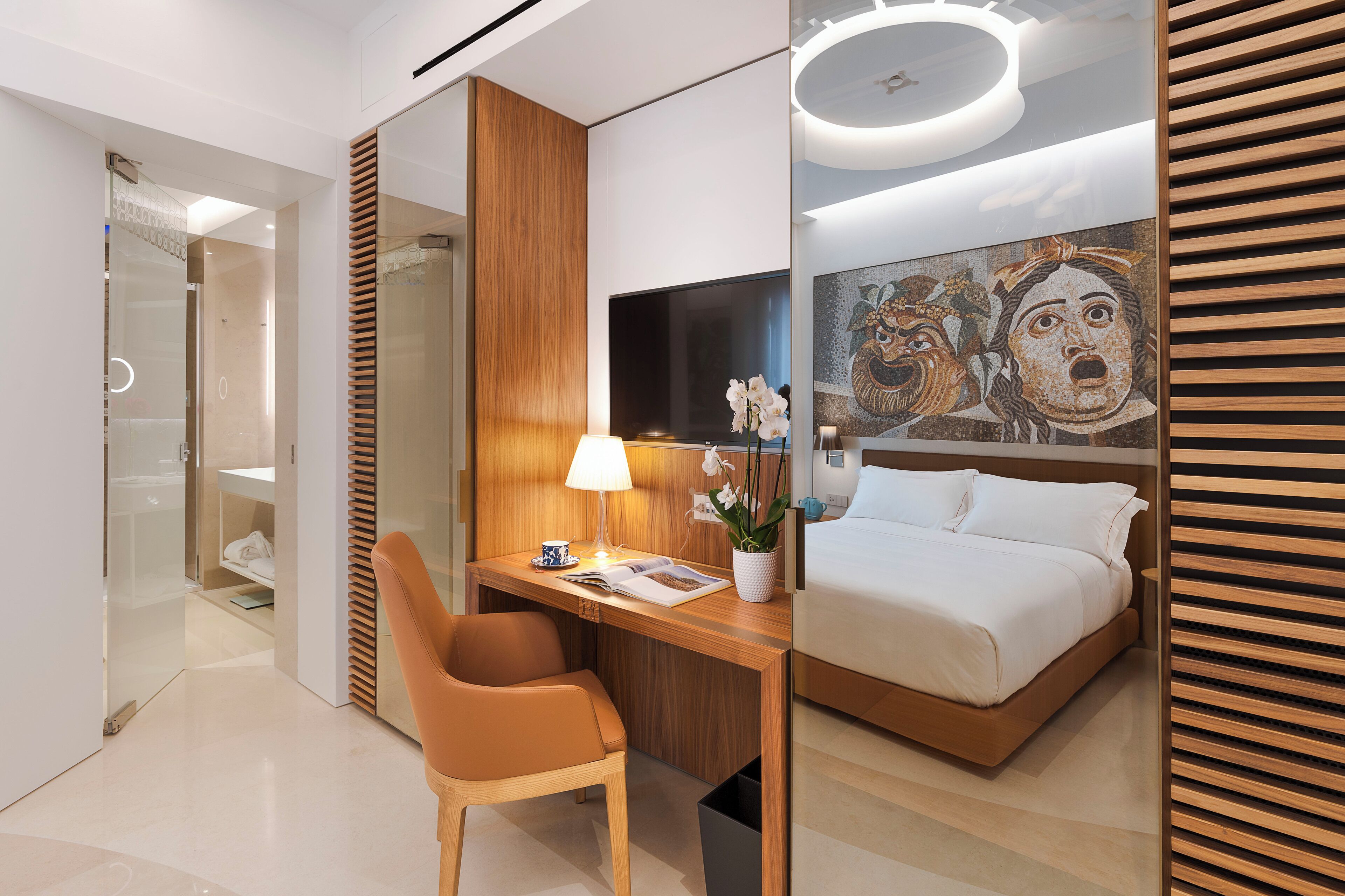 Una camera d'hotel moderna con una scrivania in legno, una poltrona confortevole, un letto con biancheria bianca, una grande TV a schermo piatto e un quadro decorativo appeso al muro.