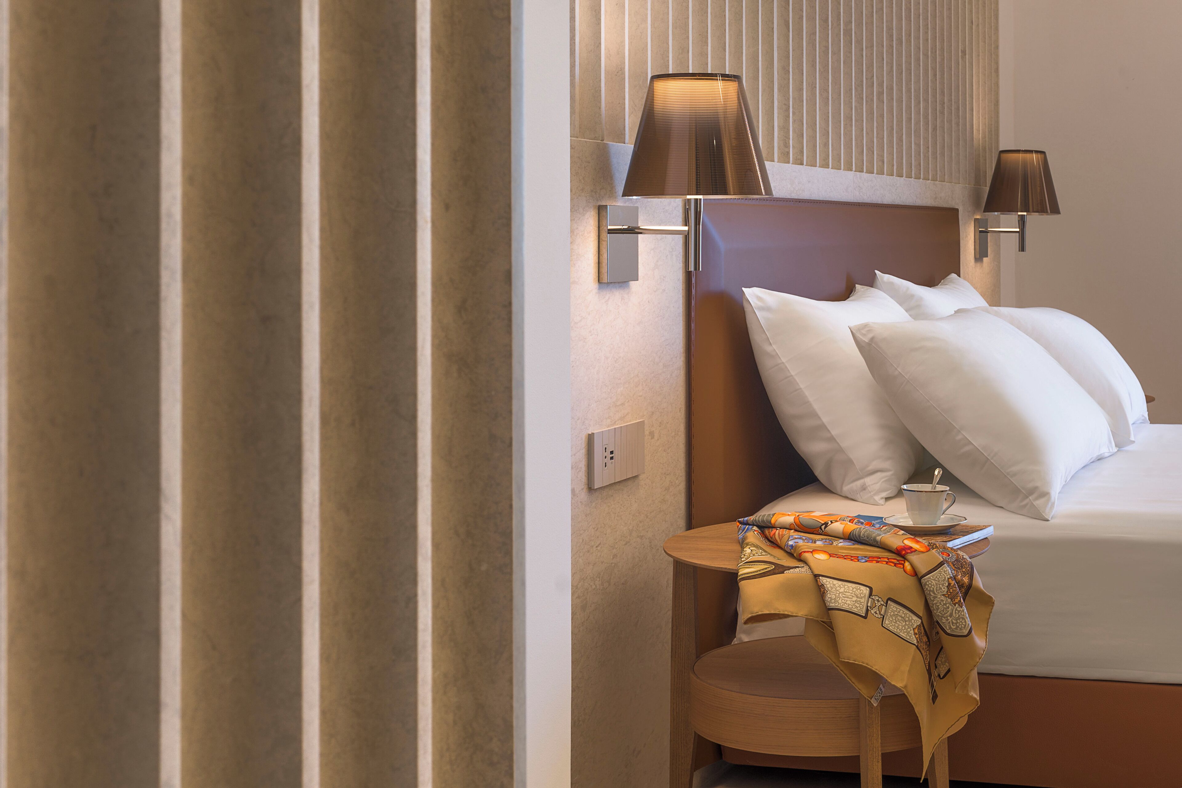 Una camera d'albergo con un letto, due lampade a parete, un comodino con tazza e piattino, e un tessuto decorativo sul tavolo.