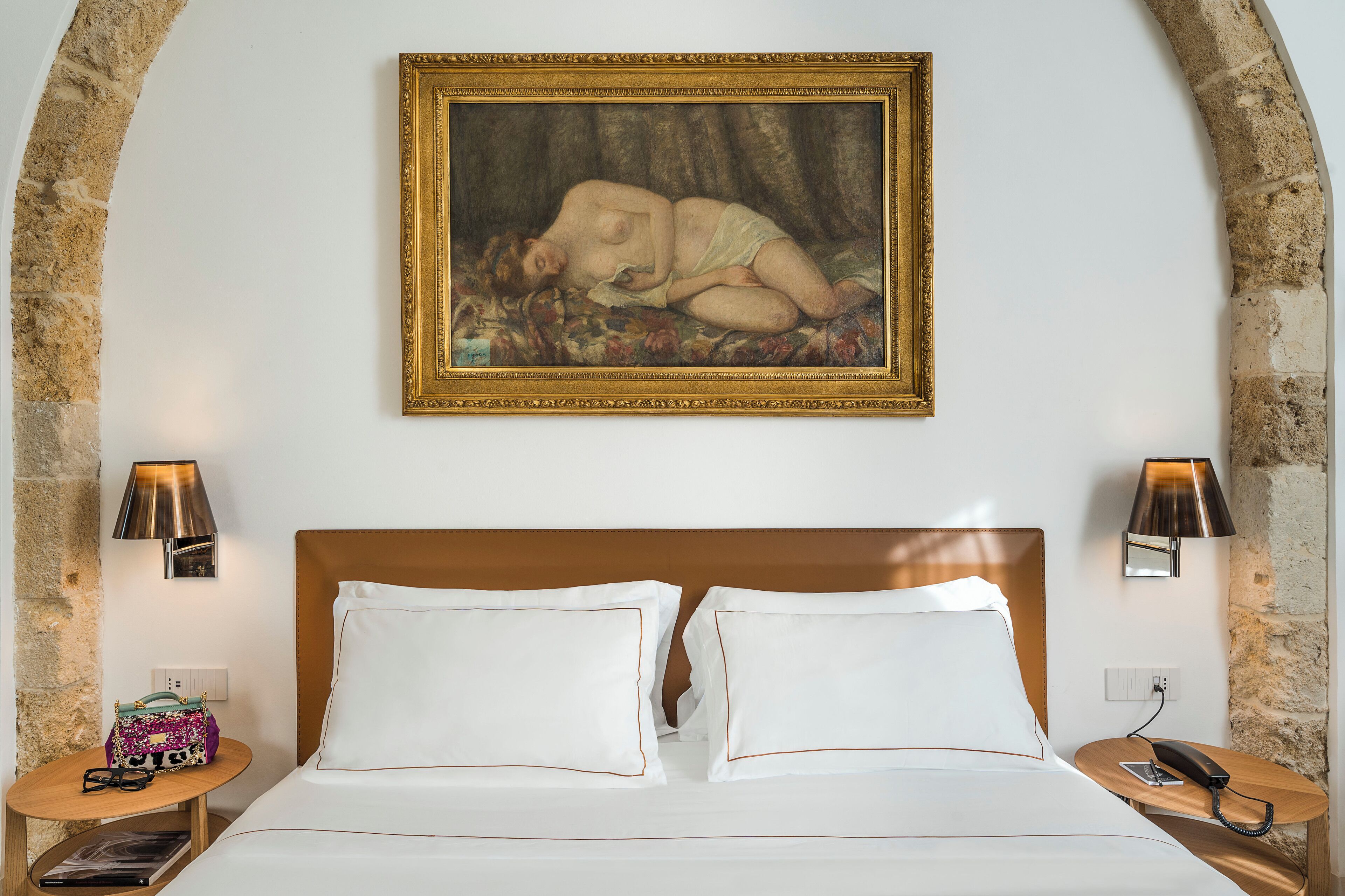 A hotel room with a bed, two bedside lamps, a painting, and a small table with a handbag and glasses.