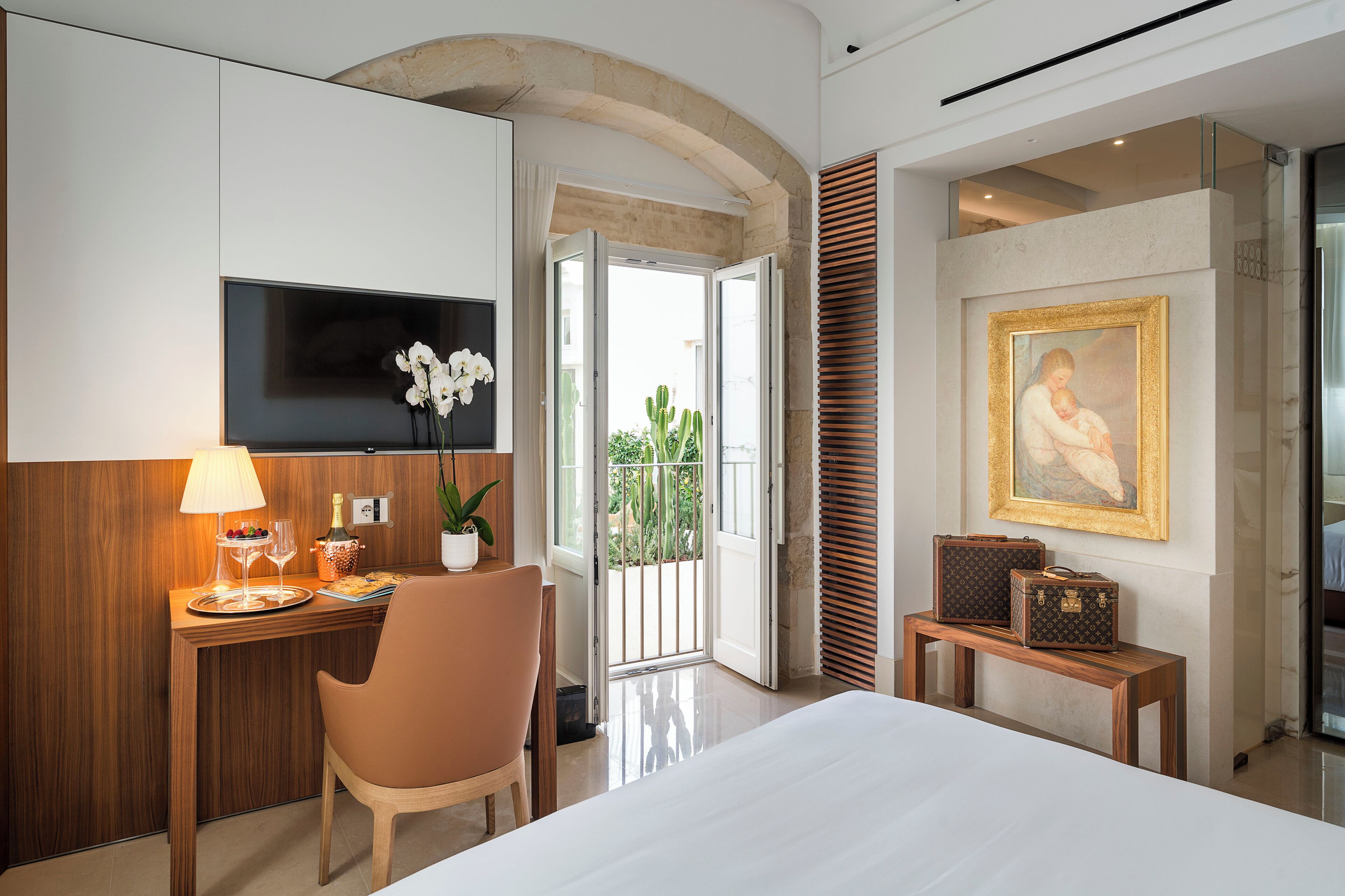 A hotel room with a bed, a desk with a lamp, a TV, a chair, a small table with suitcases, a painting, and a view of the outdoors.