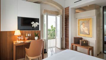 A hotel room with a bed, a desk with a lamp, a TV, a chair, a small table with suitcases, a painting, and a view of the outdoors.