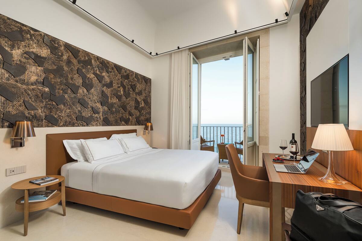 A modern hotel room with a large bed, a desk with a laptop, a chair, and a view of the sea through a balcony.