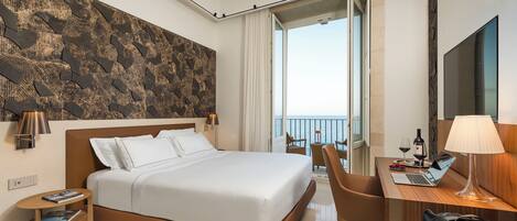Habitación de hotel moderna con una cama grande, un escritorio con una computadora portátil, una silla y vista al mar desde un balcón.