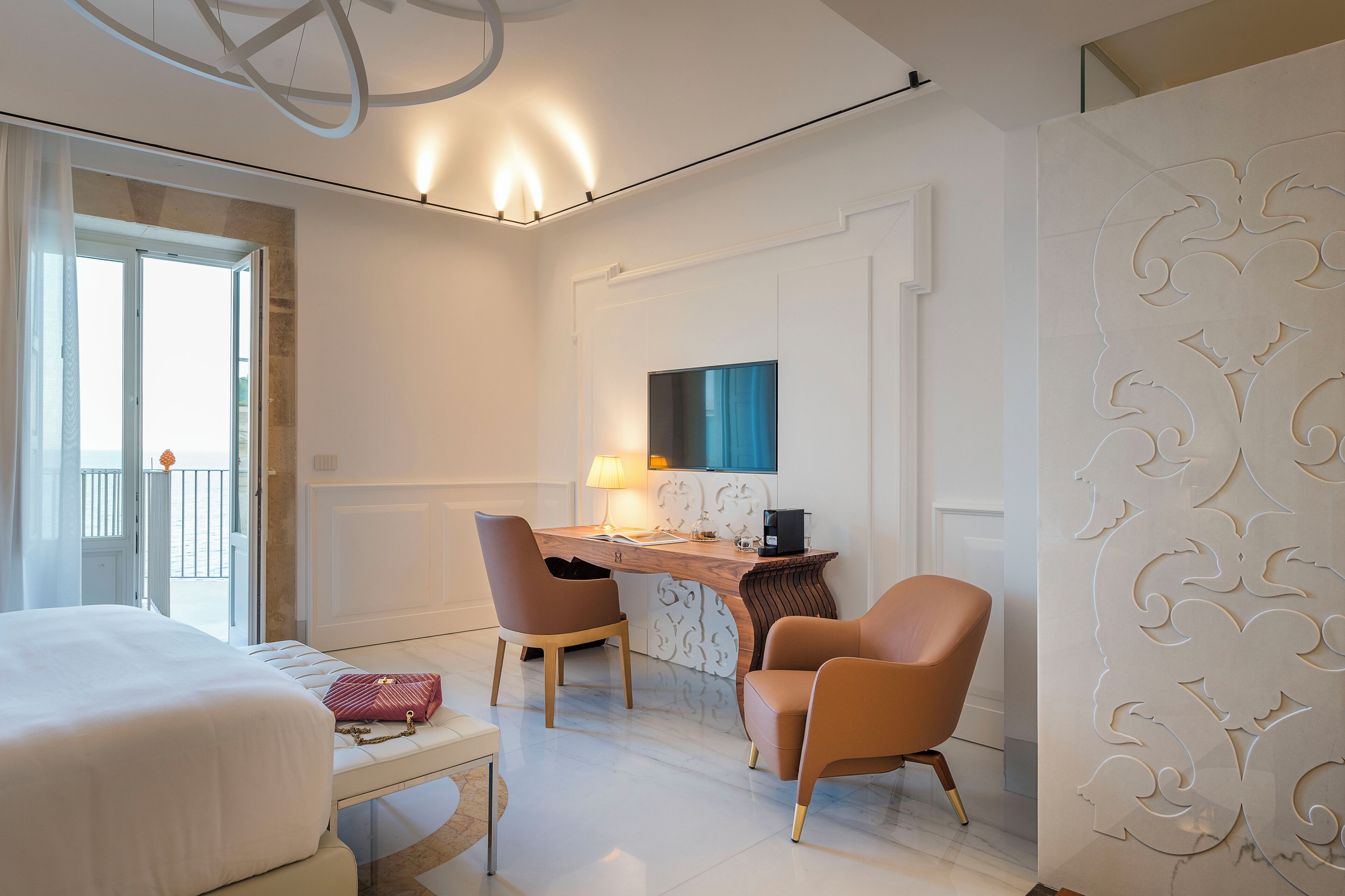A hotel room with a bed, a desk with a lamp, two chairs, and a decorative wall panel.