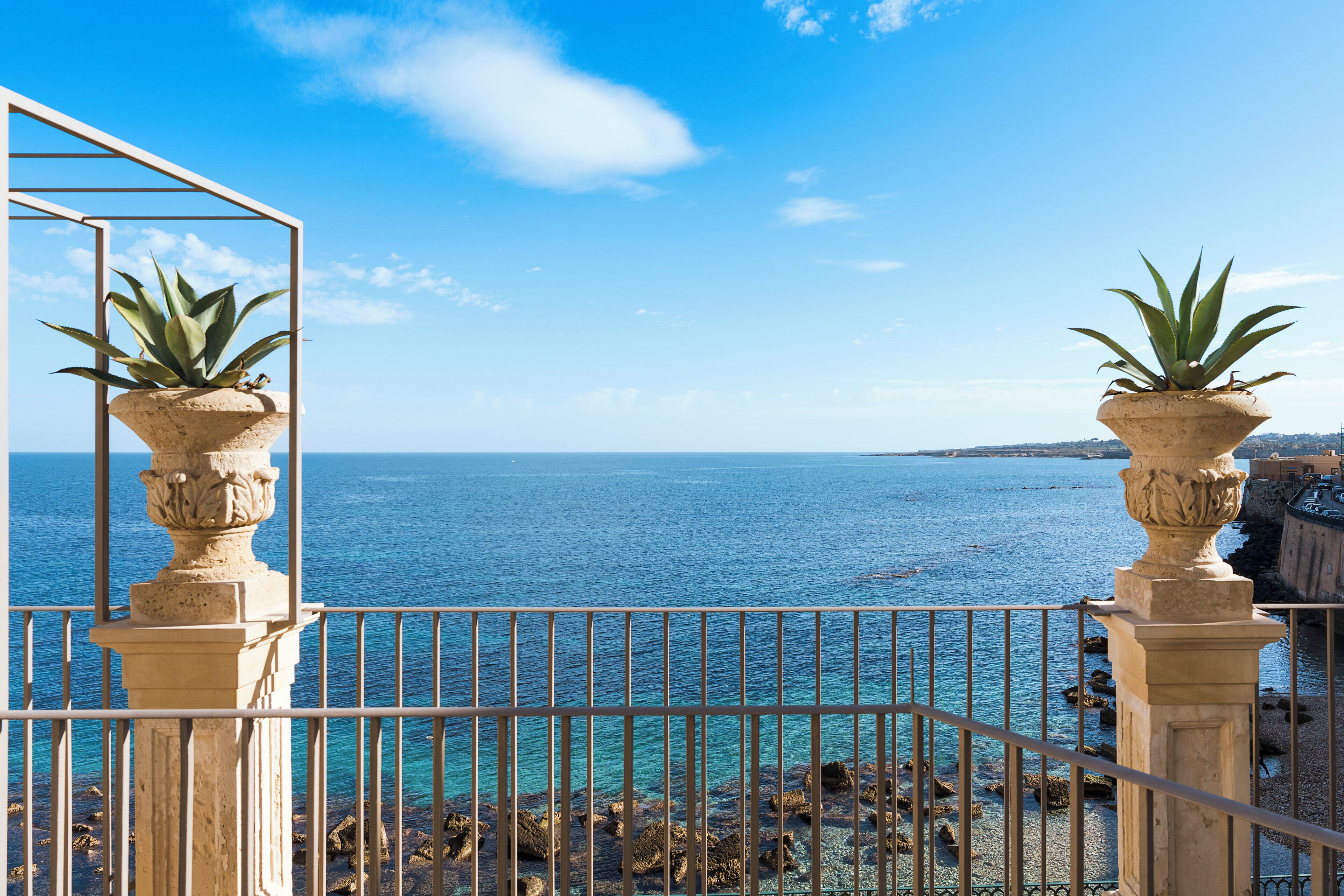 A balcony with a view of the sea, featuring two potted plants and a metal railing.