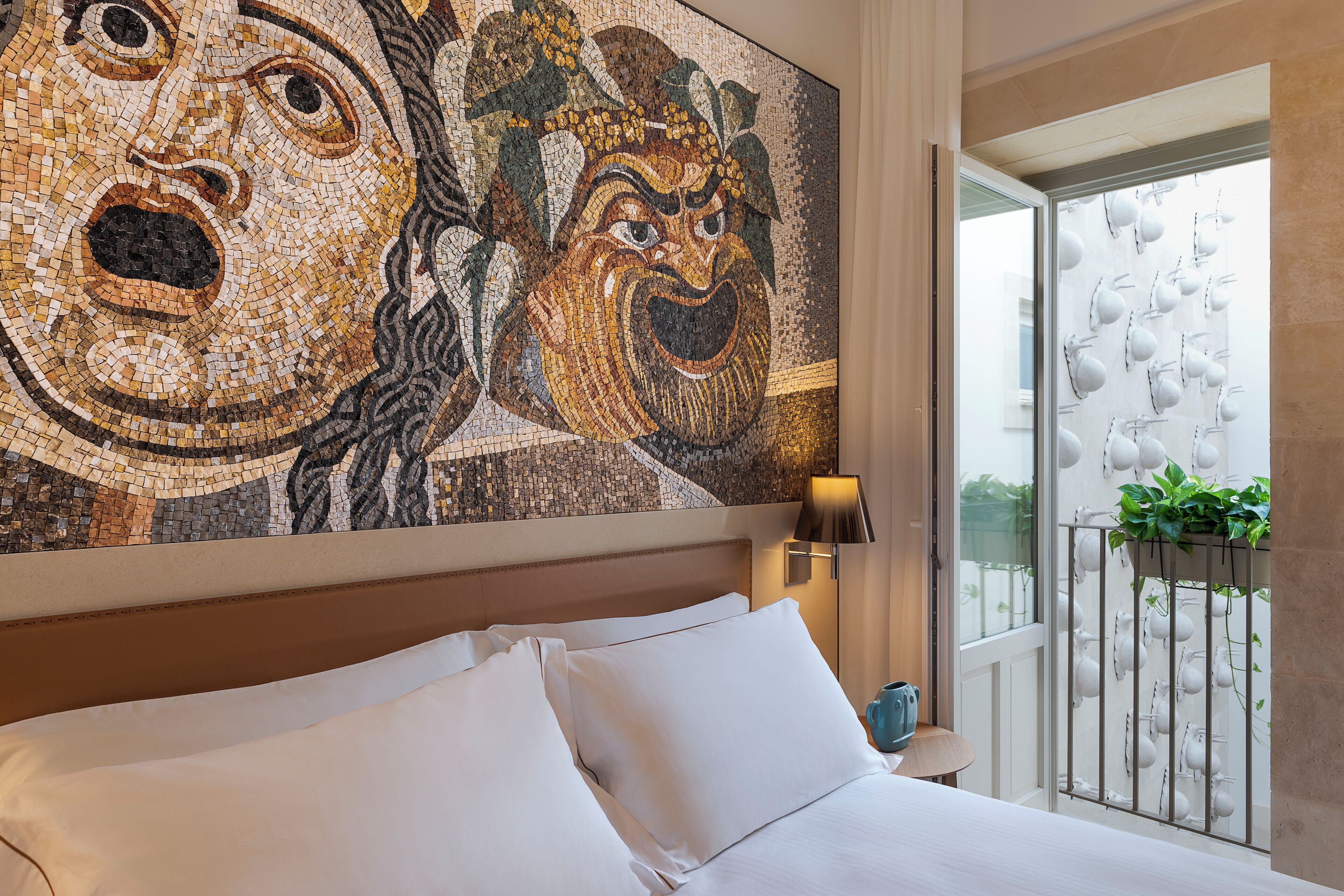 Een hotelkamer met een groot mozaiek kunstwerk aan de muur, een bed met witte lakens, een nachtkastje met een lamp en een balkon met een hekwerk en een bloempot.
